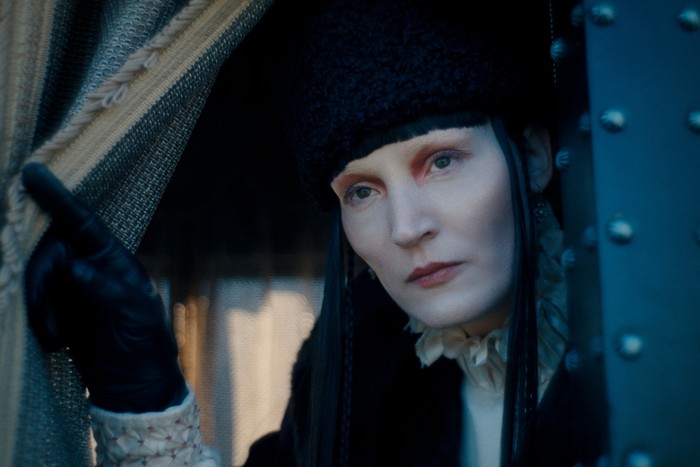 "Person in historical costume with fur hat and gloves looking out from behind a curtain in a dimly lit, possibly vintage or period setting, with serious expression and ornate clothing visible."