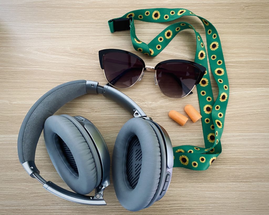 Horizontal image with grey noise-cancelling headphones, orange ear plugs, green sunflower lanyard, and black sunglasses on yellow wooden background. 