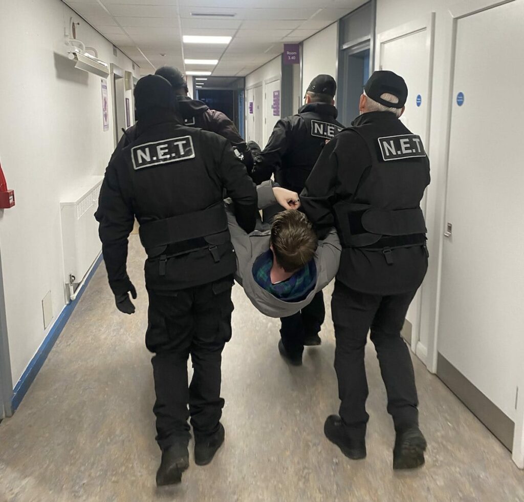 Student is carried down a corridor by baliffs