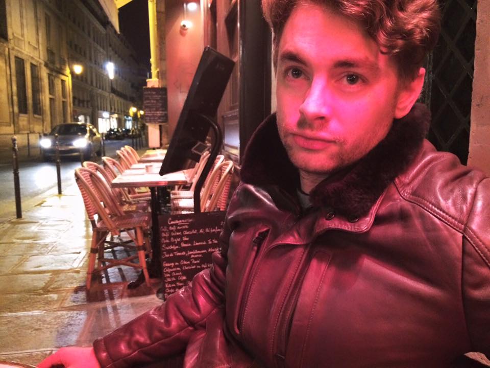 At night on a European Street, Paul Joseph Watson sits outside a restaurant. A red light is cast onto his face by the nearby signage