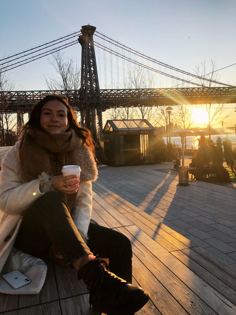 Emma Bates is sat down on what looks like a wooden pier, with a hot drink, wearing a bit winter coat and scarf as the sun sets over a bridge behind her.