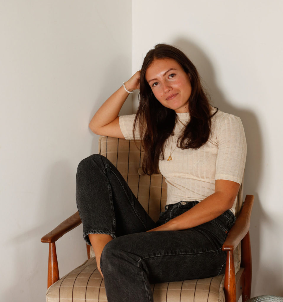 Emma Bates sits in a tartan chair, with legs crossed and her head casually resting on her hand. She wears black stonewashed jeans and a ribbed cream t-shirt.