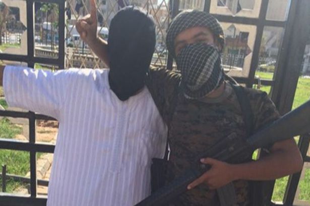 Reyhaad Khan poses next to a prisoner tied to a railing, in a tweeted picture