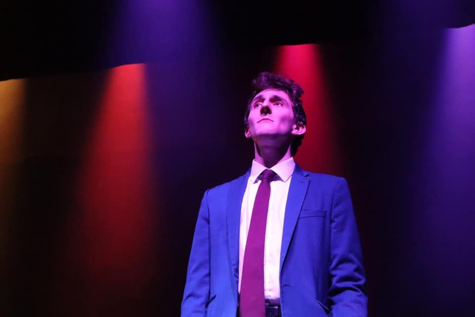 Image may contain: LED, Spotlight, Stage, Blazer, Jacket, Necktie, Suit, Apparel, Clothing, Coat, Overcoat, Human, Person, Lighting, Tie, Accessory, Accessories