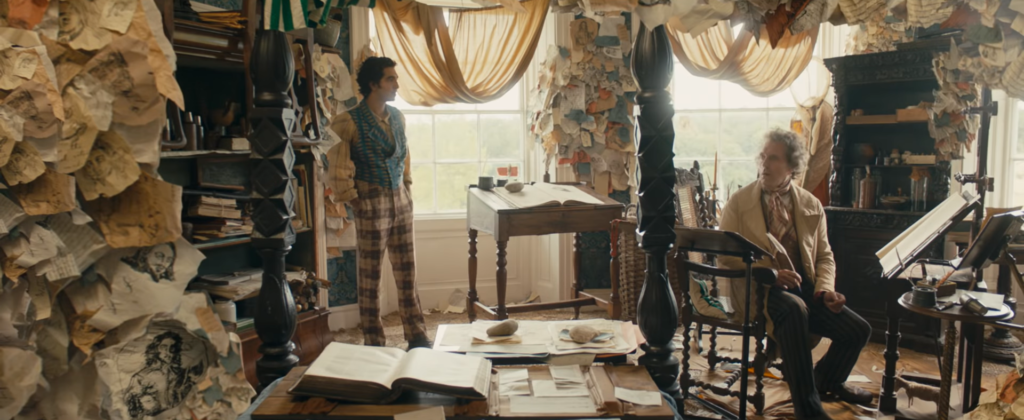 A screenshot from the movie A Personal History of David Copperfield. Two men are in a bright room which has crumpled paper all over the walls.