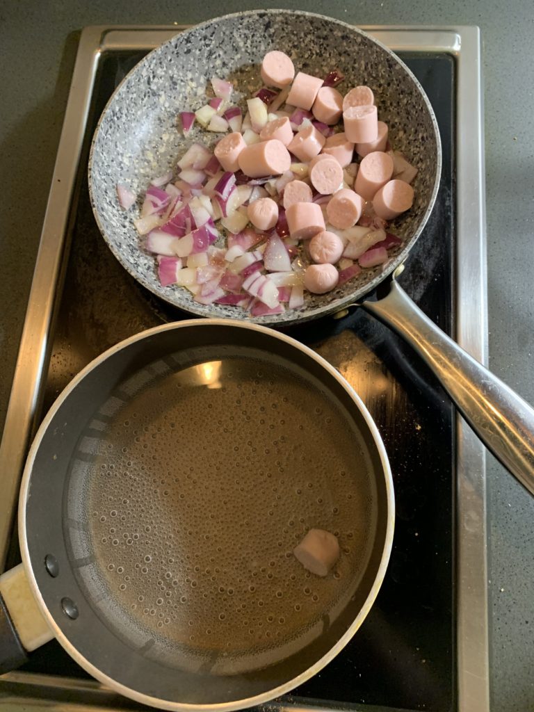 Frying pan containing chopped sausages and onion. Pot filled with water containing one slice of sausage