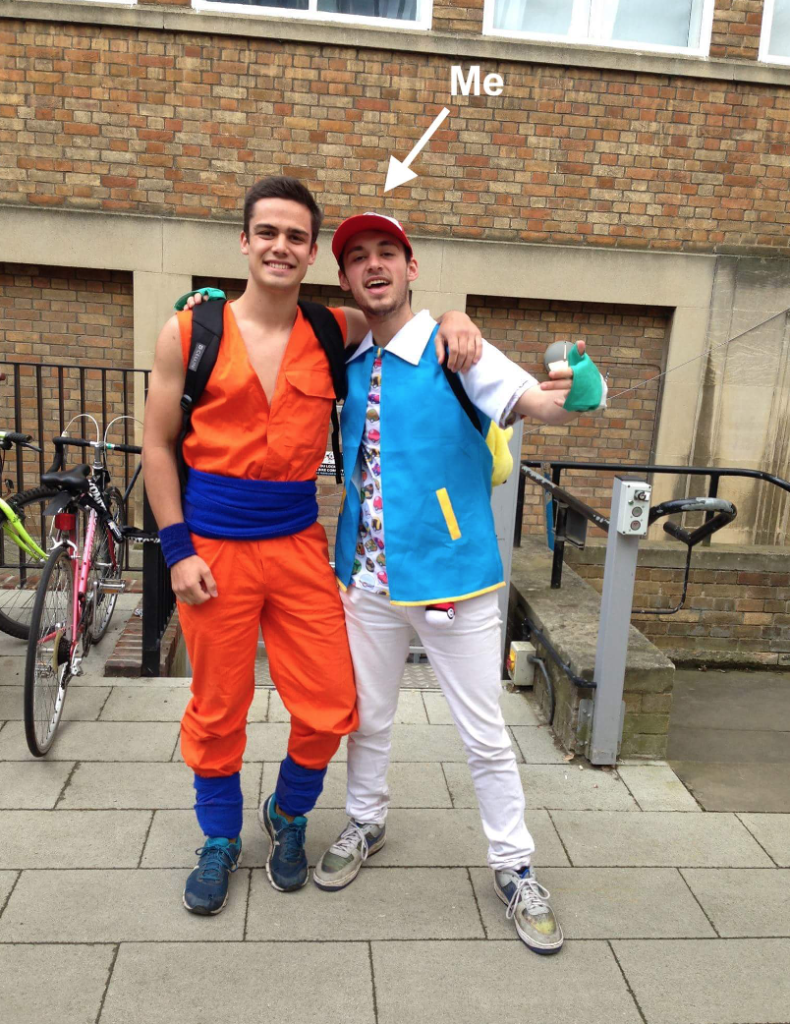 Two students hugging in new museums site near the bicycle racks. On the left, a guy is dressed as Goku from Dragon ball Z in a bright orange jumpsuit with navy bands around their waist, wrists and ankles. On the right, a guy is dressed as Ash Ketchum from Pokémon in a blue jacket, green gloves, a red cap and poke balls covering their T shirt. A white arrow labelled with the text 'Me' points to the guy dressed as Ash Ketchum on the right