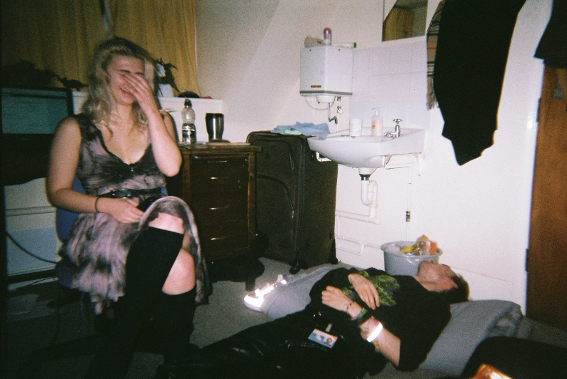 inside a college undergraduate student room. An sink with a tiny mirror can be seen next to battered furniture that looks like it's been kept since the 17th century. Two students are in the room. On the floor on the right, a man wearing black clothes and reflective bracelets is passed out, with his arms over his stomach. The man's student ID hangs from his belt. On the left, a woman is sitting above the man. The woman is wearing a cloudy black and grey dress with long black stockings. She is covering her face and laughing.