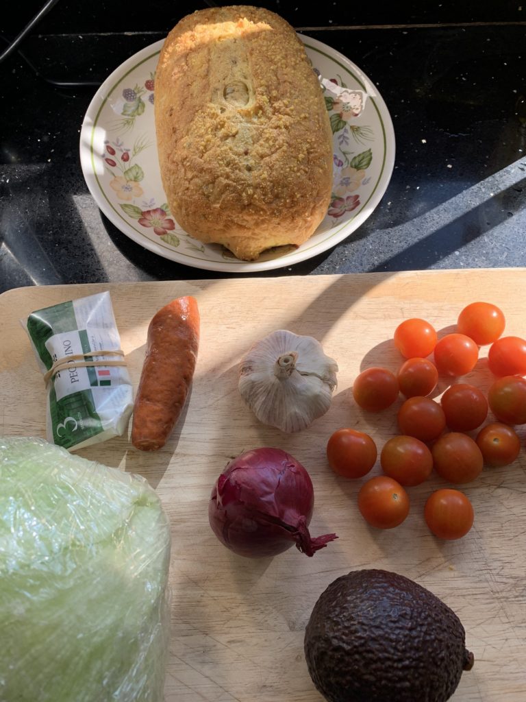 Chopping board with chorizo, cheese, cherry tomatoes, garlic, onion, avocado, lettuce and bread