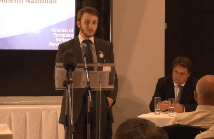 Buckby stands at a podium addressing a conference, behind him Nick Griffin sits at a table