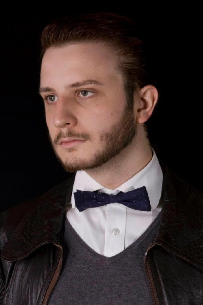 In a professional photo, Jack Buckby looks forlornly into the distance. He wears a shirt, jumper and bow tie underneath a leather bomber jacket